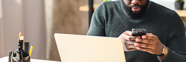 Man sms't op een smartphone terwijl hij aan een bureau zit met zijn laptop open.